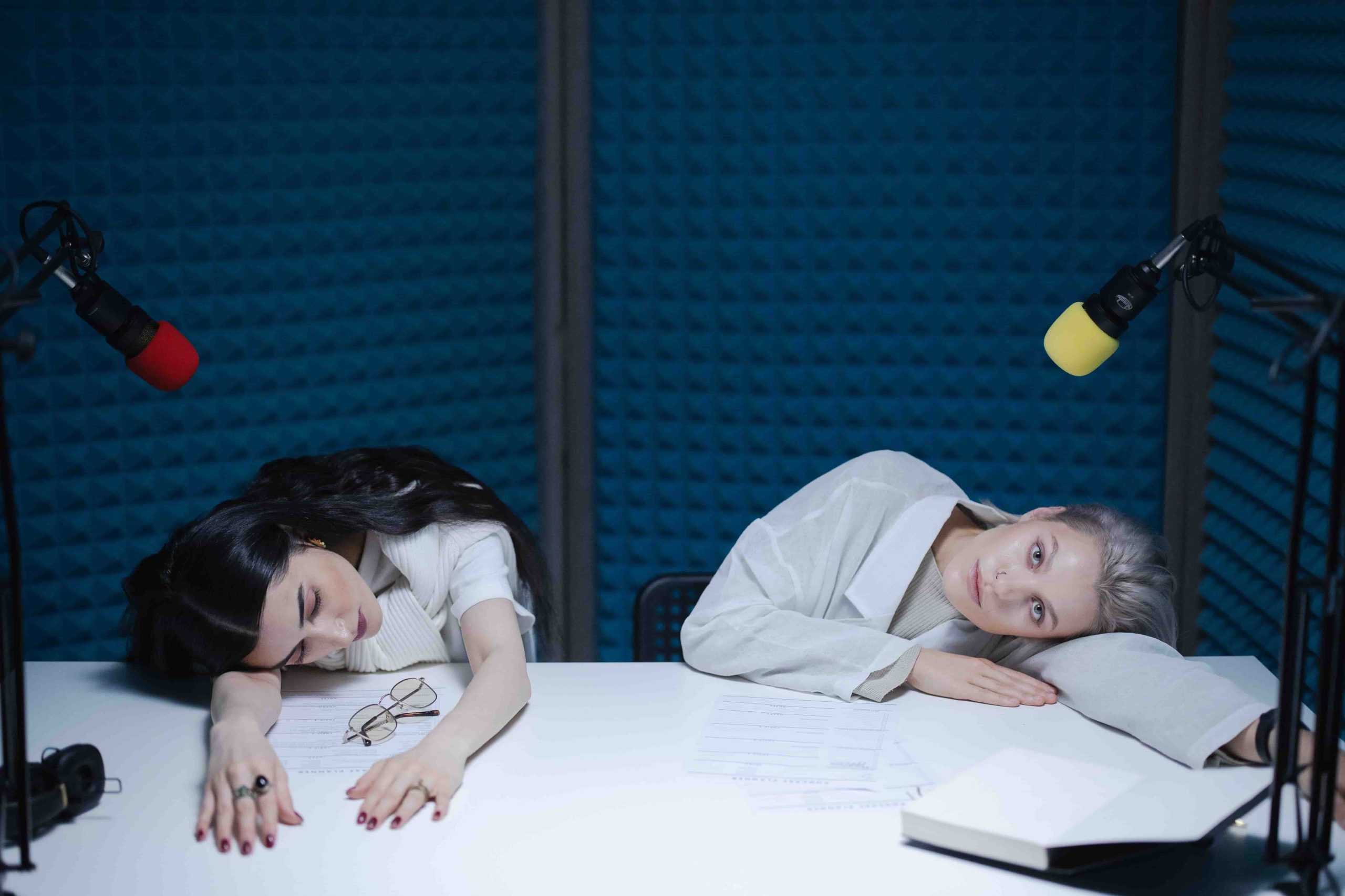 Das Bild zeigt 2 Frauen, die sich fragen, ob sie eine Podcast Produktion beauftragen sollen. Sie sind in einem Podcast Studio und scheinen müde zu sein.